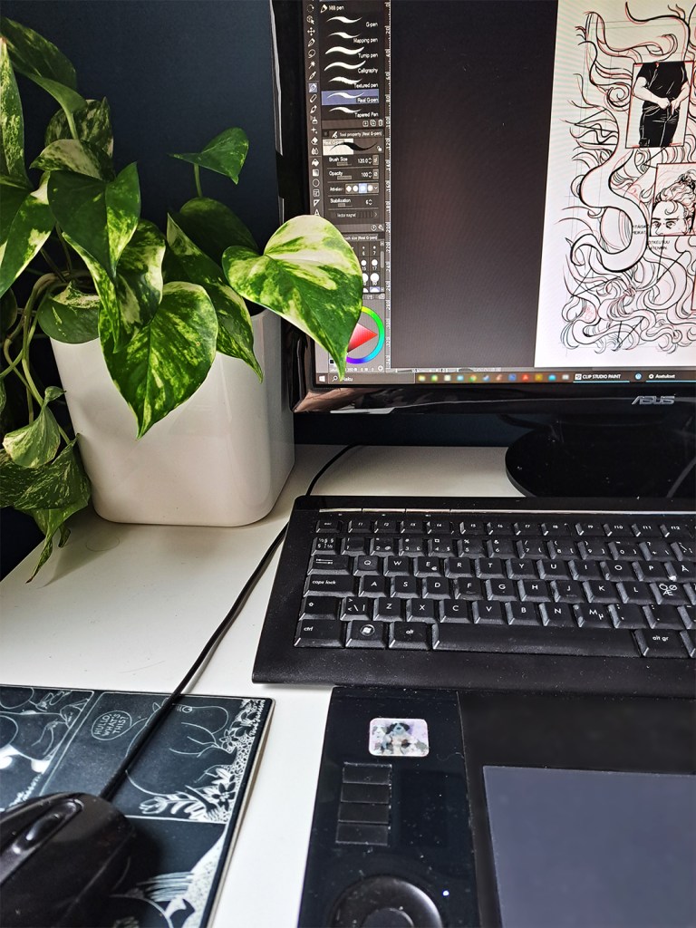 A photo of the artist's desk with a houseplant and desktop monitor, keyboard, Wacom drawing tablet and mouse. The monitor shows a draft of a comics page.